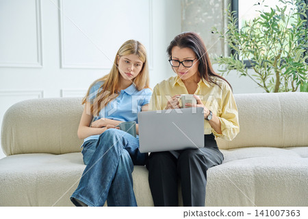 Mother and teenage daughter looking at laptop screen together on sofa Mother and teenage daughter looking at laptop screen together on sofa 141007363