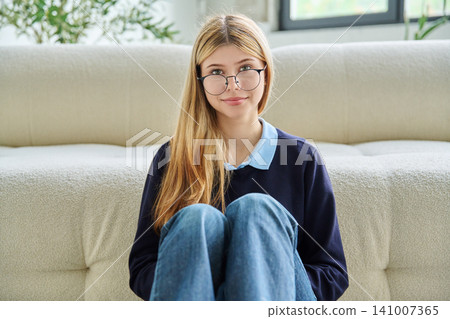 Smiling teenage student girl in glasses sitting on floor hugging knees. Smiling teenage student girl in glasses sitting on floor hugging knees. 141007365