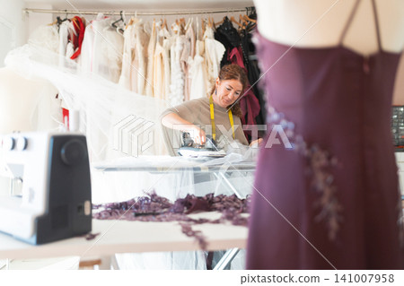 Craftswoman designer tailor working on a dress with an iron at a sewing table, surrounded by wedding gowns and fabric in a well-lit studio environment Craftswoman designer tailor working on a dress with an iron at a sewing table, surrounded by wedding gowns and fabric in a well-lit studio environment 141007958