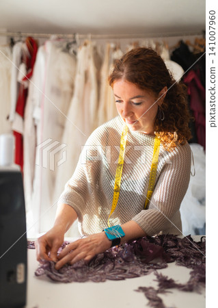 Craftswoman designer tailor working with purple fabric on a table, surrounded by various garments hanging in a well-lit sewing studio environment Craftswoman designer tailor working with purple fabric on a table, surrounded by various garments hanging in a well-lit sewing studio environment 141007960