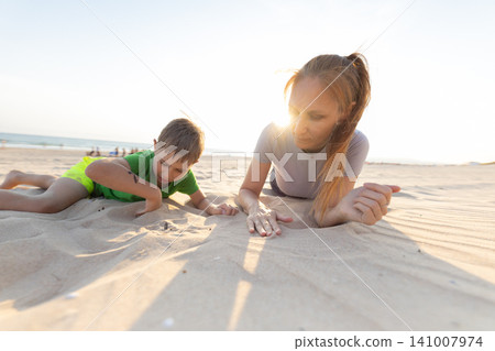 Woman and boy playing together on sandy beach, examining shells and sand, with ocean waves and sun setting in the background creating a warm atmosphere Woman and boy playing together on sandy beach, examining shells and sand, with ocean waves and sun setting in the background creating a warm atmosphere 141007974