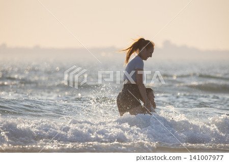 Woman in a white shirt and black shorts plays with a child in the ocean waves during sunset, creating splashes of water against a soft, blurred background Woman in a white shirt and black shorts plays with a child in the ocean waves during sunset, creating splashes of water against a soft, blurred background 141007977