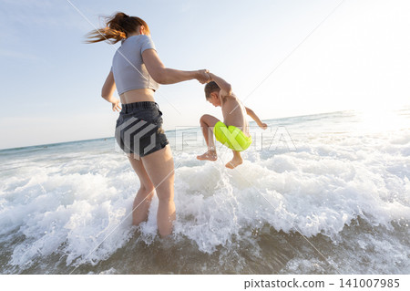 Woman in gray shirt and shorts holds boy's hand as he jumps into ocean waves during sunny day at beach, water splashing around them Woman in gray shirt and shorts holds boy's hand as he jumps into ocean waves during sunny day at beach, water splashing around them 141007985