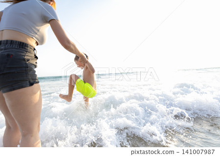 Woman in gray top and black shorts holds boy in yellow swim trunks as he jumps into ocean waves on sunny beach with bright sky and water splashes Woman in gray top and black shorts holds boy in yellow swim trunks as he jumps into ocean waves on sunny beach with bright sky and water splashes 141007987