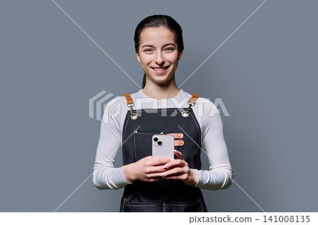 Confident young female in apron using smartphone posing on gray background Confident young female in apron using smartphone posing on gray background 141008135