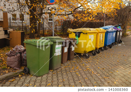 recycling bins stationary in an outdoor urban area with trash bags on a sidewalk and fallen leaves at ground level, ordinary scene with green and yellow tones, environmental conservation. recycling bins stationary in an outdoor urban area with trash bags on a sidewalk and fallen leaves at ground level, ordinary scene with green and yellow tones, environmental conservation. 141008360