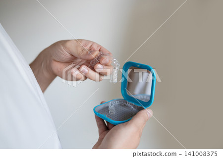 dentist holding and displaying clear aligners in a blue plastic case against a neutral background in a close-up hand-held perspective, professional and highly informative. dentist holding and displaying clear aligners in a blue plastic case against a neutral background in a close-up hand-held perspective, professional and highly informative. 141008375