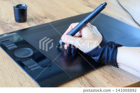 Designer hand in glove drawing with stylus pen on graphic tablet, digital art creation process on wooden table closeup Designer hand in glove drawing with stylus pen on graphic tablet, digital art creation process on wooden table closeup 141008892