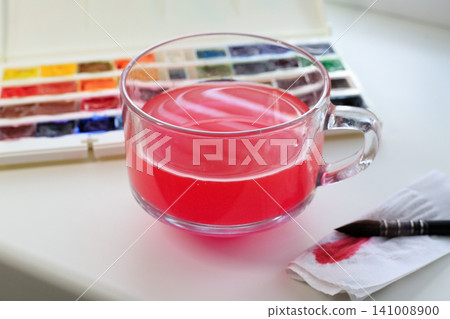 Workspace of a watercolor artist with a glass of water mixed with pink paint, a brush, a stained napkin, and a palette with watercolor paints on a white table. Workspace of a watercolor artist with a glass of water mixed with pink paint, a brush, a stained napkin, and a palette with watercolor paints on a white table. 141008900