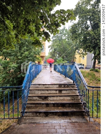 A young woman with a red umbrella walks across a bridge on a rainy day. 141009437