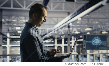 Data center engineer monitors operational metrics on laptop inside a server room to improve infrastructure performance. Data center engineer monitors operational metrics on laptop inside a server room to improve infrastructure performance. 141009839