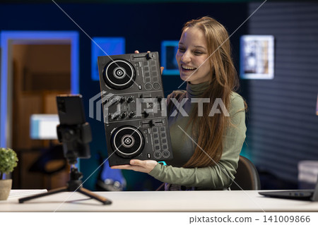 Young girl influencer reviewing DJ controller board at home, demonstrating mixing equipment with knobs and soundboard features. Content creator testing electronic console functions on video. Young girl influencer reviewing DJ controller board at home, demonstrating mixing equipment with knobs and soundboard features. Content creator testing electronic console functions on video. 141009866