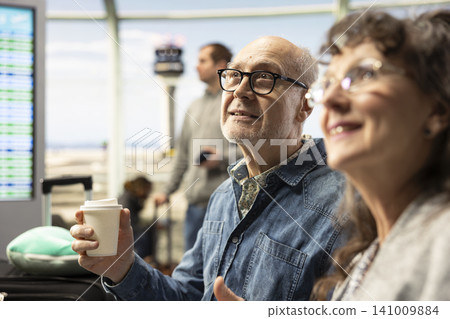 Smiling elderly man and woman, drinking coffee, in airport lounge area, discussing ideas and travel plans before flight. Confident retirees representing active lifestyle and fulfilling life goals. Smiling elderly man and woman, drinking coffee, in airport lounge area, discussing ideas and travel plans before flight. Confident retirees representing active lifestyle and fulfilling life goals. 141009884
