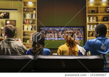 Close up of fans watching football match on large television screen at home. Back view of supporters waiting in anticipation for favorite team to score during soccer game Close up of fans watching football match on large television screen at home. Back view of supporters waiting in anticipation for favorite team to score during soccer game 141009887