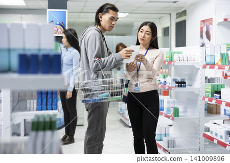 Portrait of man holding shopping basket with prescription slip, talking with pharmacy employee. Young male adult browsing and discussing products in aisle filled with health and wellness items. Portrait of man holding shopping basket with prescription slip, talking with pharmacy employee. Young male adult browsing and discussing products in aisle filled with health and wellness items. 141009896