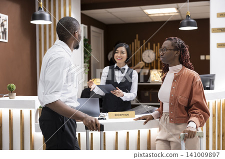 Hotel concierge welcoming travelers couple at reception counter of resort lobby. Guests with luggage complete the registration and check-in process while hotel staff provides customer service. 141009897