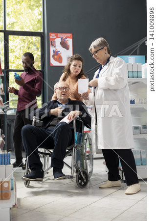 Caucasian woman pharmacist in lab coat assists senior customer in wheelchair at local drugstore. Medical specialist discussing prescription medication and suitable health products in calm consultation Caucasian woman pharmacist in lab coat assists senior customer in wheelchair at local drugstore. Medical specialist discussing prescription medication and suitable health products in calm consultation 141009898