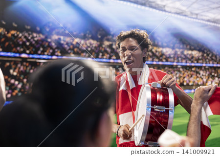 Excited football supporter beats drum among cheering fans in packed stadium during soccer match day. Fan leader creating rhythm with musical instrument, creating loud crowd energy chant Excited football supporter beats drum among cheering fans in packed stadium during soccer match day. Fan leader creating rhythm with musical instrument, creating loud crowd energy chant 141009921