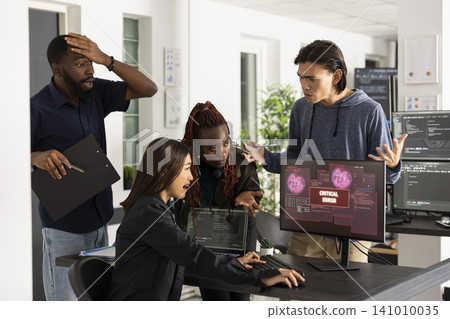 Professional IT team expresses shock and urgency while facing major cybersecurity attack. Frustrated developers looking at desktop monitor displaying red critical error warnings during data breach. Professional IT team expresses shock and urgency while facing major cybersecurity attack. Frustrated developers looking at desktop monitor displaying red critical error warnings during data breach. 141010035