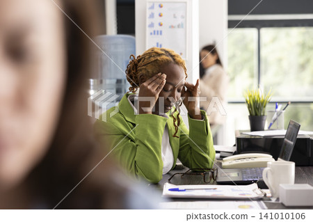 African American woman leans forward at desk with eyes closed, dealing with pounding tension headache. Female manager feels fatigue and emotional strain from constant deadlines in corporate office. African American woman leans forward at desk with eyes closed, dealing with pounding tension headache. Female manager feels fatigue and emotional strain from constant deadlines in corporate office. 141010106
