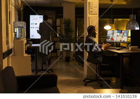 African american male, online videocall, office desk, working late after hours, man focused on monitor to lead briefing presentation and engagement with colleagues across time zones through chat. 141010139