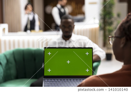 Green screen laptop in hotel lounge area with tourists couple, relaxing on couch in lobby during early arrival. Hospitality industry environment shows guests awaiting check-in. Green screen laptop in hotel lounge area with tourists couple, relaxing on couch in lobby during early arrival. Hospitality industry environment shows guests awaiting check-in. 141010140