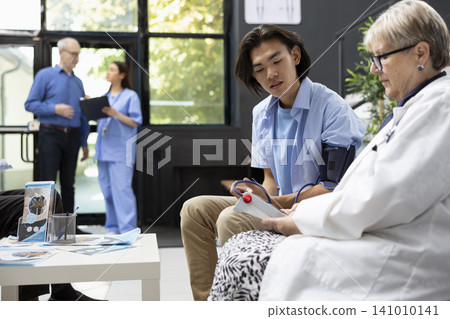 Asian man wears arm blood pressure cuff in hospital lobby while female doctor analyzes vital signs on monitoring device. Physician conducts routine checkup of male patient prior to medical examination Asian man wears arm blood pressure cuff in hospital lobby while female doctor analyzes vital signs on monitoring device. Physician conducts routine checkup of male patient prior to medical examination 141010141