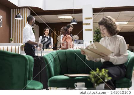Hotel concierge assisting tourists at the hotel front desk upon arrival. Asian woman staff provides reservation confirmation and registration support while guests wait in the stylish lobby. Hotel concierge assisting tourists at the hotel front desk upon arrival. Asian woman staff provides reservation confirmation and registration support while guests wait in the stylish lobby. 141010160