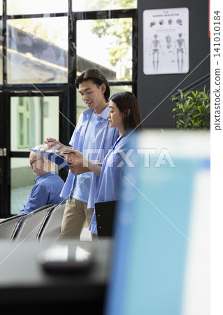 Female healthcare worker in scrubs guides patient through health insurance form at hospital. Nurse carefully reviews information on clipboard, ensuring accuracy before upcoming medical consultation. Female healthcare worker in scrubs guides patient through health insurance form at hospital. Nurse carefully reviews information on clipboard, ensuring accuracy before upcoming medical consultation. 141010184