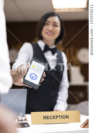 Tourist making contactless payment with NFC on smartphone at hotel service desk, POS terminal completes transaction while hotel staff finalize check-in forms. Elegant reception area at resort. Tourist making contactless payment with NFC on smartphone at hotel service desk, POS terminal completes transaction while hotel staff finalize check-in forms. Elegant reception area at resort. 141010211