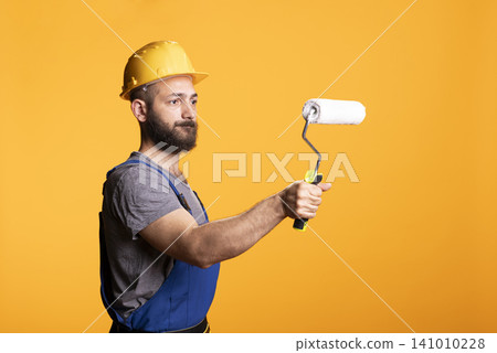 Painter in hardhat demonstrates roller for decorating or renovation jobs, presenting handyman service concept. Male construction worker stands in studio with painting tool against yellow backdrop Painter in hardhat demonstrates roller for decorating or renovation jobs, presenting handyman service concept. Male construction worker stands in studio with painting tool against yellow backdrop 141010228