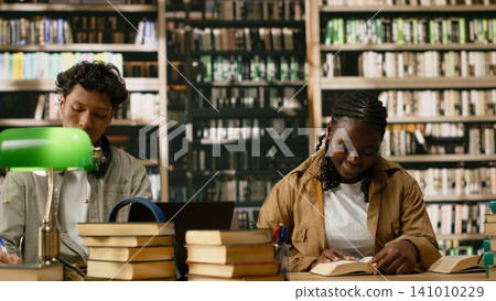 Black woman student reading open book at shared library table, study area with soft focus on shelves in quiet space. Lecture in study session in academic workspace. Black woman student reading open book at shared library table, study area with soft focus on shelves in quiet space. Lecture in study session in academic workspace. 141010229