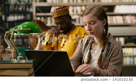 Student typing on laptop and taking notes beside open book at busy study table, study group with stacks of books and green lamps. Young woman writing on keyboard in library. 141010230