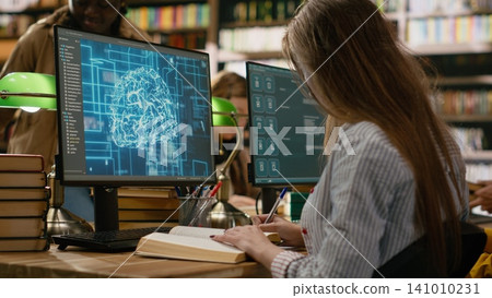 Student research AI brain visuals on desktop computer while reading books and taking notes at shared library technology desk for digital learning. Computer class with science. Student research AI brain visuals on desktop computer while reading books and taking notes at shared library technology desk for digital learning. Computer class with science. 141010231