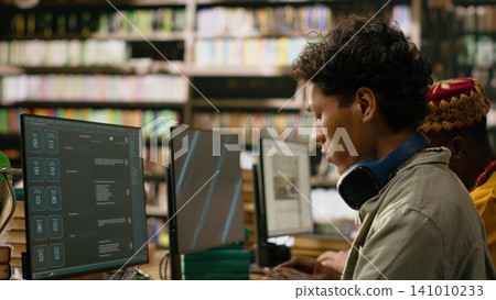 Asian student reading open book beside desktop monitor and keyboard while classmates work at nearby computers in warm library study environment. STEM education and learning. Asian student reading open book beside desktop monitor and keyboard while classmates work at nearby computers in warm library study environment. STEM education and learning. 141010233