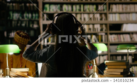 Back view of young woman seated with classmates in dark library study room, surrounded by green lamps and open books. Tall bookshelves for quiet academic research. 141010234
