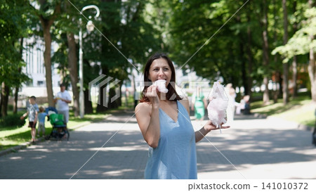 A woman enjoys eating cotton candy in a relaxed and pleasant park pathway surrounded by trees and people, capturing a summer fun family outing with vibrant blue and lush. A woman enjoys eating cotton candy in a relaxed and pleasant park pathway surrounded by trees and people, capturing a summer fun family outing with vibrant blue and lush. 141010372