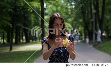 Woman eating ice cream, in park, with pathway, trees, people in the background, with paper cup with colorful, wooden stick for ice texture, from a frontal view perspective. Woman eating ice cream, in park, with pathway, trees, people in the background, with paper cup with colorful, wooden stick for ice texture, from a frontal view perspective. 141010373