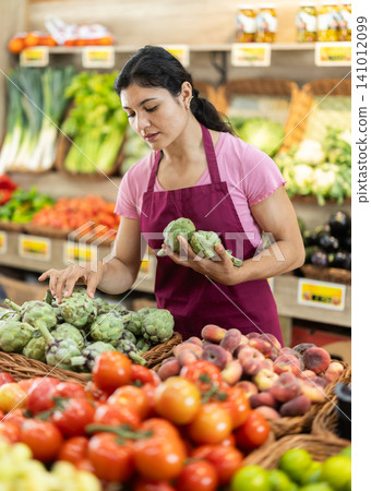 Adult woman seller puts artichokes in vegetable shop Adult woman seller puts artichokes in vegetable shop 141012099