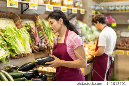 Female seller wearing apron holding zucchini and eggplants in hand in grocery shop Female seller wearing apron holding zucchini and eggplants in hand in grocery shop 141012201