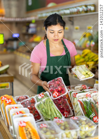 Female salesperson places vegetables and fruits packed in plastic containers on supermarket display case. 141012257