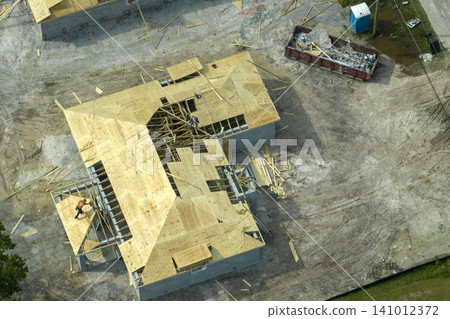 Professional builders working on roof construction of unfinished suburban home with wooden frame structure in Florida rural area. Concept of housing development in America Professional builders working on roof construction of unfinished suburban home with wooden frame structure in Florida rural area. Concept of housing development in America 141012372