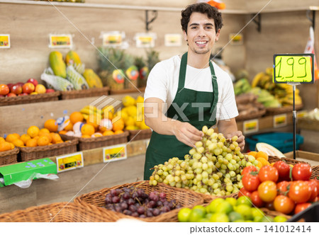 Male seller wearing apron holding grape in hand in grocery shop Male seller wearing apron holding grape in hand in grocery shop 141012414