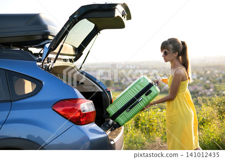 Young woman in summer dress putting green suitcase inside her car trunk. Travel and vacations concept. Young woman in summer dress putting green suitcase inside her car trunk. Travel and vacations concept. 141012435