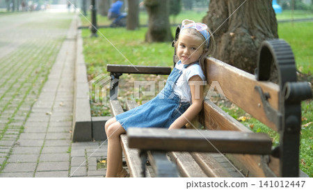 Small happy child girl sitting on a bench resting in summer park. Small happy child girl sitting on a bench resting in summer park. 141012447