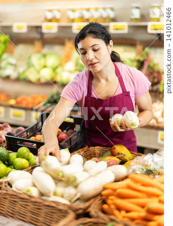 Adult woman seller puts daikon in vegetable shop Adult woman seller puts daikon in vegetable shop 141012466