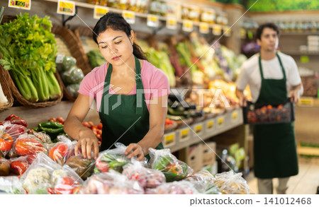 Female salesperson places vegetables packed in plastic bags on supermarket display case. Female salesperson places vegetables packed in plastic bags on supermarket display case. 141012468