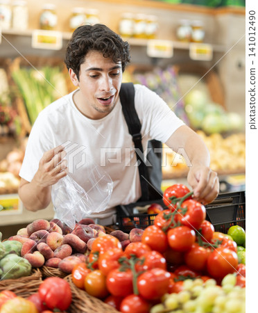 Man buys ripe tomatoes in a supermarket. European woman stands near a vegetable counter and holds tomatoes in her hands, examines them and chooses them to cook dinner Man buys ripe tomatoes in a supermarket. European woman stands near a vegetable counter and holds tomatoes in her hands, examines them and chooses them to cook dinner 141012490