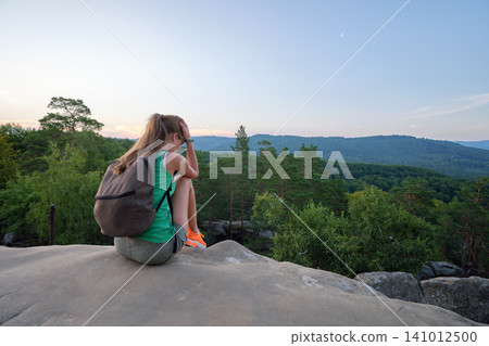 Young sad woman hiker sitting alone on rocky mountain on warm evening. Solitude and resting from everyday rush concept Young sad woman hiker sitting alone on rocky mountain on warm evening. Solitude and resting from everyday rush concept 141012500