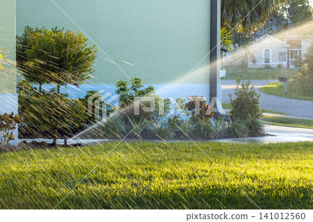Sprinkler irrigating grass lawn with water in summer garden. Watering green vegetation during dry season for maintaining it fresh Sprinkler irrigating grass lawn with water in summer garden. Watering green vegetation during dry season for maintaining it fresh 141012560